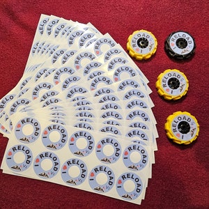 May include: A stack of white stickers with the text "I ♥ RELOAD V2" printed in black and blue. There are four yellow and black plastic caps with the same text printed on them.