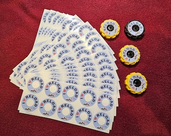 Puede incluir: Una pila de pegatinas blancas con el texto "I ♥ RELOAD V2" impreso en negro y azul. Hay cuatro tapas de plástico amarillo y negro con el mismo texto impreso en ellas.