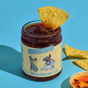 May include: A jar of Salsa Macha with a tortilla chip on top, plus more chips and a lid, on a blue background. The jar label has illustrations and text. The jar contains a dark red sauce.