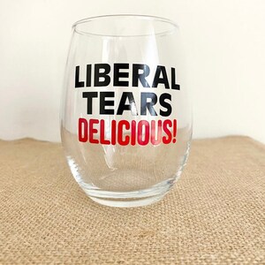May include: A clear glass wine glass with the text "LIBERAL TEARS DELICIOUS!" printed on it in black and red.