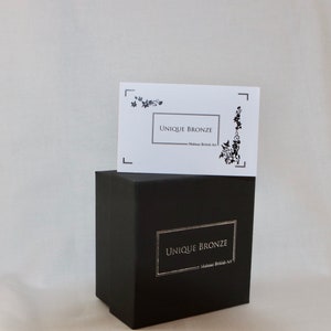 Puede incluir: Una caja negra con una etiqueta blanca que dice "UNIQUE BRONZE - Made in British Art". La caja est&aacute; sobre una superficie blanca.