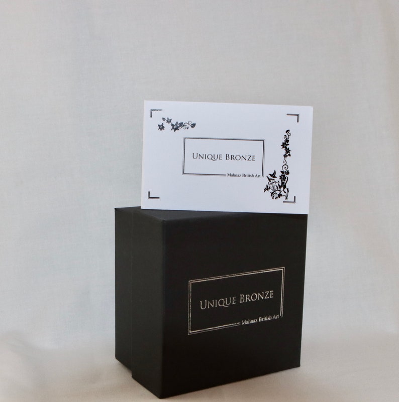 Puede incluir: Una caja negra con una etiqueta blanca que dice "UNIQUE BRONZE - Made in British Art". La caja est&aacute; sobre una superficie blanca.