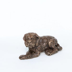 May include: A bronze-colored sculpture of a puppy in a resting pose. The puppy is lying down with its front paws extended and head raised. The sculpture has a textured surface and detailed features.