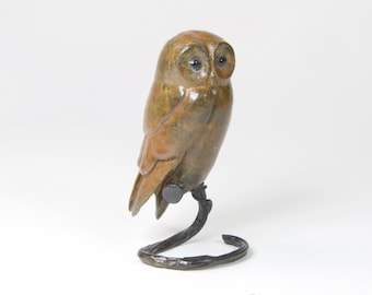 May include: A bronze sculpture of an owl perched on a metal branch. The owl has brown and green feathers and large, round eyes.