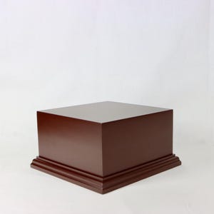 May include: A dark brown wooden display stand with a square top and a decorative base. The stand is made of solid wood and has a smooth, glossy finish. The base has a layered design.