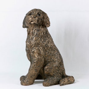 Pode incluir: Uma escultura de bronze de um cão sentado com a cabeça levantada. O cão tem uma pelagem encaracolada e uma expressão doce.