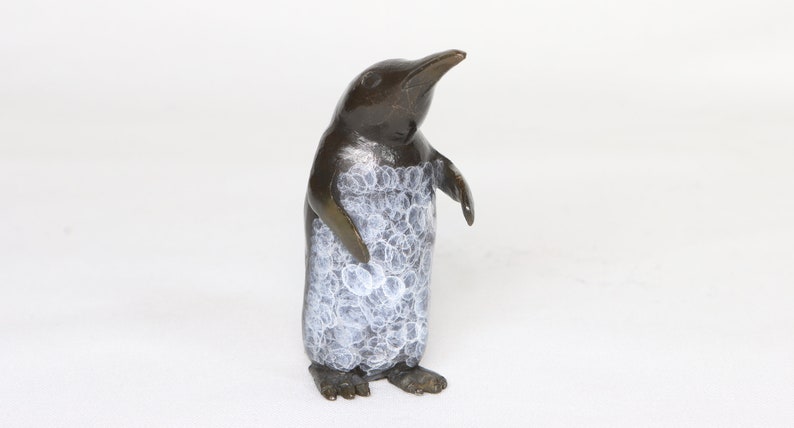 Puede incluir: Una peque&ntilde;a escultura de ping&uuml;ino de bronce con un dise&ntilde;o pintado de blanco en su cuerpo. El ping&uuml;ino est&aacute; de pie con la cabeza inclinada hacia arriba.