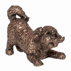 May include: A bronze sculpture of a dog in a playful pose. The dog is lying on its belly with its head turned to the side and its tail curled up. The sculpture is made in a textured style, giving the dog a furry appearance.