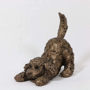 May include: A bronze-colored sculpture of a dog in a playful pose, with its front paws extended and tail raised. The dog's fur appears textured, and the sculpture is set against a plain white background.