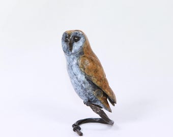 May include: A bronze-colored owl sculpture with a light blue and brown finish. The owl is perched on a curved branch, with its head turned slightly to the side. The sculpture is set against a white background.