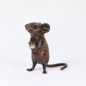 Peut inclure: Une figurine de souris en bronze, debout, les pattes jointes. La sculpture présente des détails, notamment les oreilles, les yeux et une longue queue. La souris semble tenir un petit objet rond. La figurine est de couleur bronze.