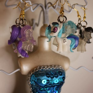 May include: Four pairs of colorful pony-themed earrings on a jewelry display stand. The earrings are made of plastic and feature different pony characters with bright colors and details.