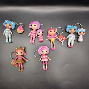 May include: A selection of doll keyrings in various colours. The dolls are styled with different hairstyles and outfits, such as pyjamas, dresses, and bear costumes. Some keyrings include additional charms like a cupcake, a bear, and a lollipop. The keyrings are attached to silver metal rings.