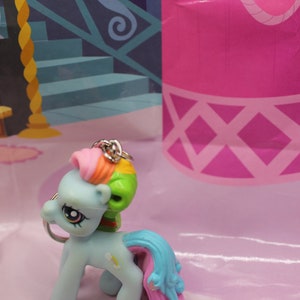May include: A light blue My Little Pony keychain with a rainbow mane and tail. The pony has a green head and a silver keyring. The background is a pink surface with a cartoon castle.
