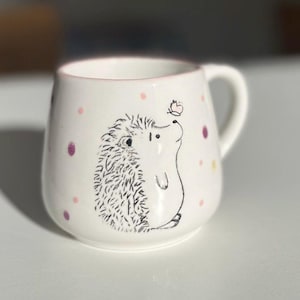 Könnte beinhalten: Weiße Keramiktasse mit rosa Rand und einer Schwarzweißillustration eines Igels, der zu einem Schmetterling aufblickt. Die Tasse hat ein rosa-gelbes Punkte-Muster.