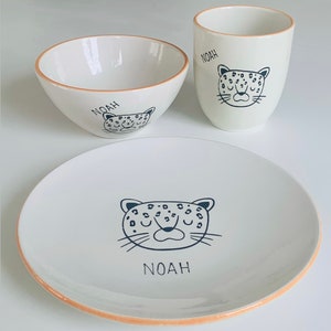 Könnte beinhalten: Ein weißes Kinder-Dinner-Set aus Keramik mit Leopardenmuster. Das Set enthält eine Schüssel, einen Teller und eine Tasse. Der Name "Noah" ist auf jedes Stück gedruckt.