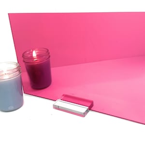 May include: A pink acrylic sheet with a reflective surface. Two candles are in front of the sheet, one is lit and the other is not.