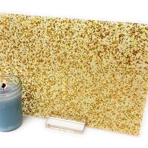 Puede incluir: Una lámina de vinilo iridiscente de purpurina dorada con una base amarillo claro. El vinilo está sobre una superficie blanca con un soporte de acrílico transparente.