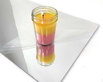 May include: A yellow and pink layered candle in a clear glass jar with a wick. The candle is sitting on a silver reflective surface.