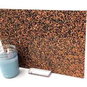 May include: A sheet of black and orange glitter paper with a speckled pattern. The paper is propped up on a white surface with a blue candle jar in the foreground.