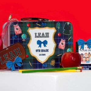 Puede incluir: Un conjunto escolar personalizado con una lonchera a cuadros, una etiqueta de equipaje y una tarjeta de fotos escolares. La lonchera y la etiqueta llevan el nombre "Leah", un lazo azul y gráficos escolares. Una manzana roja y lápices completan la escena.