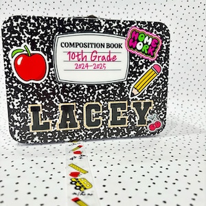 Può includere: Una lunchbox nera e bianca con un design a quaderno di composizione. Ha una mela rossa, una matita e un adesivo che dice "Home Work". La lunchbox ha anche il testo "Composition Book 10th Grade 2024-2025" e il nome "LACEY" in lettere dorate.