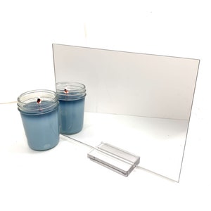 May include: A clear acrylic mirror with a beveled edge, standing on a clear acrylic stand. Two blue glass candle jars are in front of the mirror.