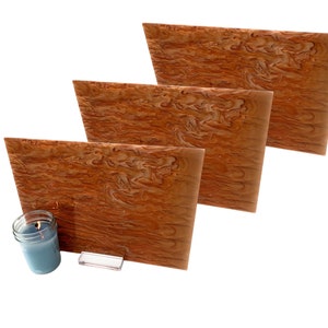 Op de afbeelding: Drie rechthoekige houten panelen met een wervelend, gemarmerd patroon in bruin- en beigetinten. Een brandende blauwe kaars in een glazen pot en een klein, helder rechthoekig object staan op de voorgrond. De panelen zijn trapsgewijs gerangschikt.