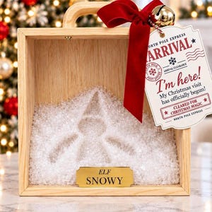 Puede incluir: Una caja de madera clara con asa, rellena de material cristalino blanco con forma de silueta de elfo. Lleva atada una cinta de terciopelo rojo con un cascabel dorado y una etiqueta blanca que dice NORTH POLE EXPRESS ARRIVAL I'm here!. Una placa dorada dice ELF SNOWY.