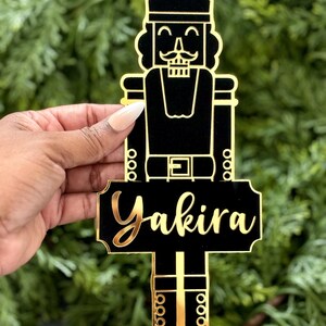 May include: A black and gold acrylic Christmas ornament in the shape of a nutcracker soldier. The ornament has the name "Yakira" in gold script. The nutcracker is wearing a crown and has a mustache.