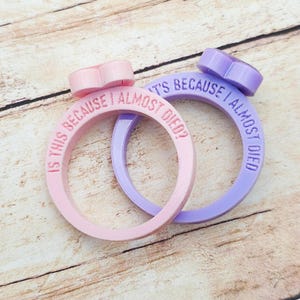 Könnte beinhalten: Zwei ringförmige Objekte, eines rosa und eines lila, mit herzförmigen Akzenten. Der rosa Ring hat den Text "IS THIS BECAUSE I ALMOST DIED?" und der lila Ring hat den Text "IT'S BECAUSE I ALMOST DIED?"