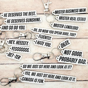 May include: A collection of white keychains with various black text phrases, including "He Deserves the Best," "Mister Business Man," and "Canada is Fun." Each keychain has a silver clasp.