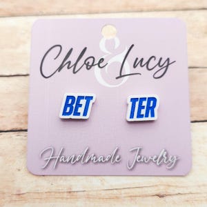 May include: A pair of white stud earrings with the words "BET" and "TER" in blue block letters. The earrings are on a light purple card with the text "Chloe & Lucy" and "Handmade Jewelry".
