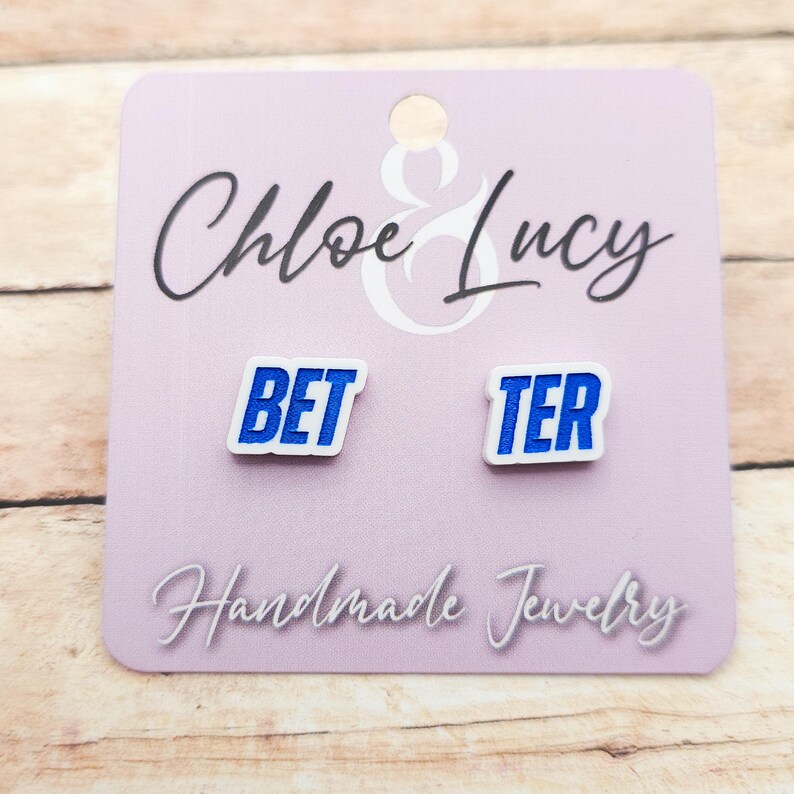 May include: A pair of white stud earrings with the words "BET" and "TER" in blue block letters. The earrings are on a light purple card with the text "Chloe & Lucy" and "Handmade Jewelry".