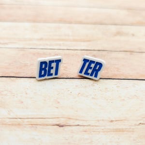 May include: Two small, white rectangular objects with the words "BET" and "TER" in blue block letters. The objects are slightly angled and set against a light brown wooden background.