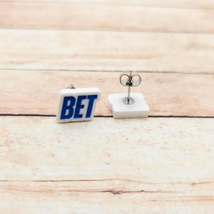 May include: A pair of square stud earrings. Each earring features the word "BET" in blue block letters on a white background. The earrings have silver posts and butterfly backs.