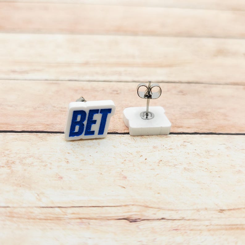 May include: A pair of square stud earrings. Each earring features the word "BET" in blue block letters on a white background. The earrings have silver posts and butterfly backs.