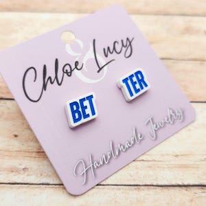 May include: A pair of white stud earrings with the words "BET" and "TER" in blue block letters. The earrings are displayed on a light purple card with the text "Chloe & Lucy Handmade Jewelry".
