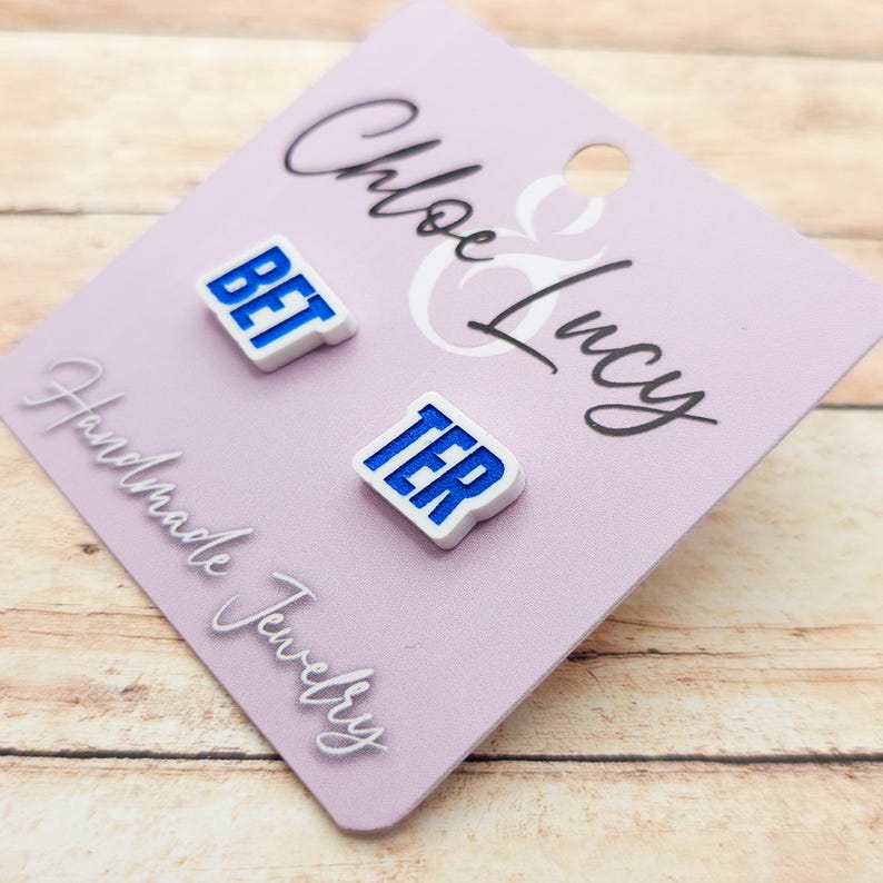 May include: A pair of white stud earrings with the words "BET" and "TER" in blue block letters. The earrings are displayed on a light purple card with the words "Chloe & Lucy Handmade Jewelry" in white script.