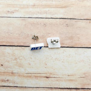 May include: White square stud earrings with the word "BET" in blue lettering. The earrings have a silver post and backing. The earrings are displayed on a light brown wooden surface.