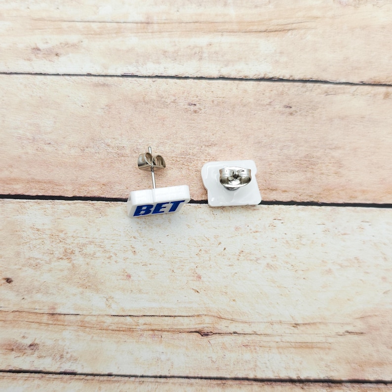 May include: White square stud earrings with the word "BET" in blue lettering. The earrings have a silver post and backing. The earrings are displayed on a light brown wooden surface.