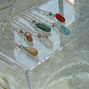 May include: Six silver necklaces with different colored gemstone pendants. The pendants are all oval shaped and are displayed on a clear acrylic surface.