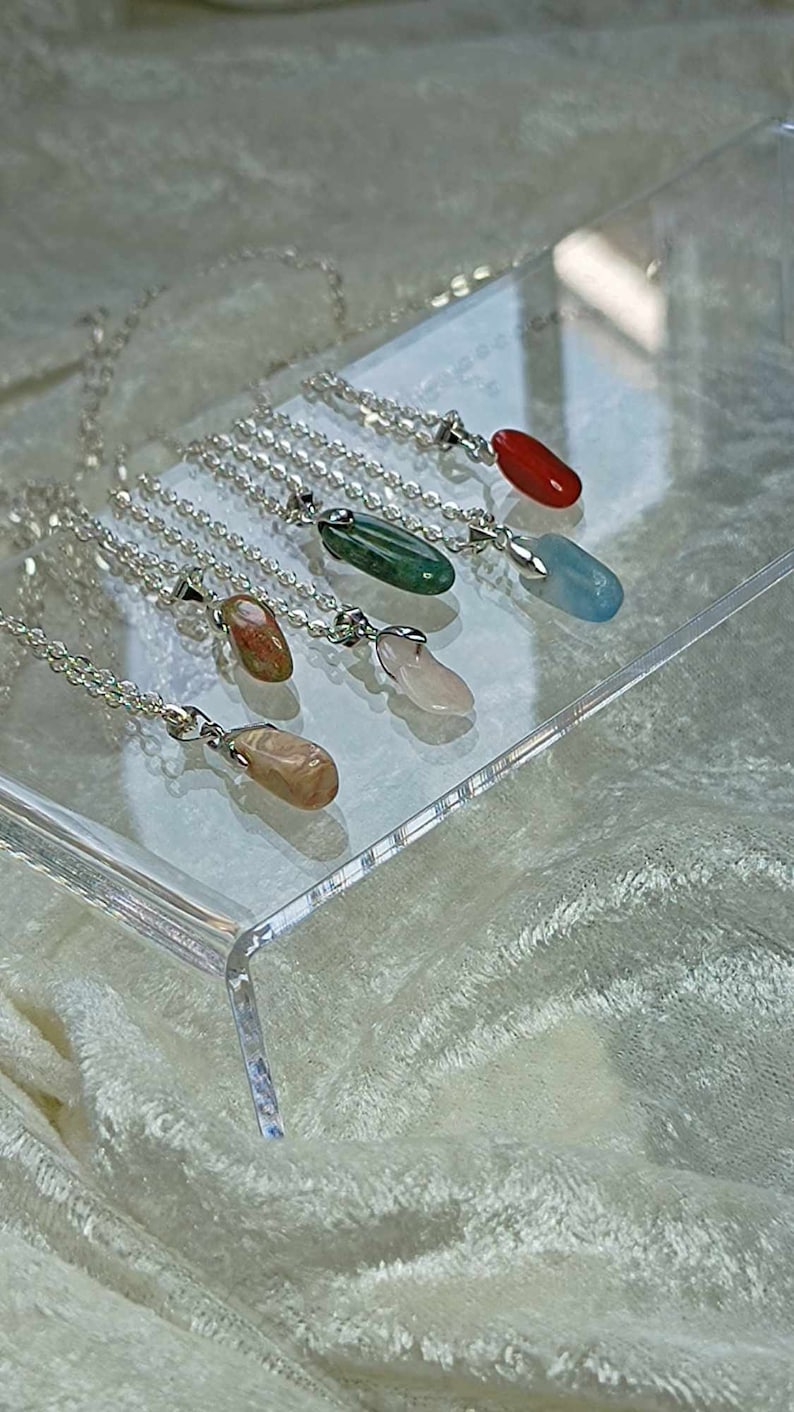 May include: Six silver necklaces with different colored gemstone pendants. The pendants are all oval shaped and are displayed on a clear acrylic surface.
