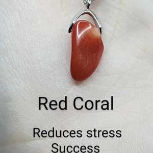 May include: Red coral pendant necklace on a silver chain. The pendant is a smooth, polished, red coral stone. The text below the pendant reads "Red Coral", "Reduces stress", "Success", and "Tranquility".
