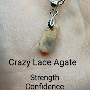 May include: A silver chain necklace with a pendant made of a light brown and white Crazy Lace Agate stone. The text "Strength Confidence Motivation" is written below the stone.