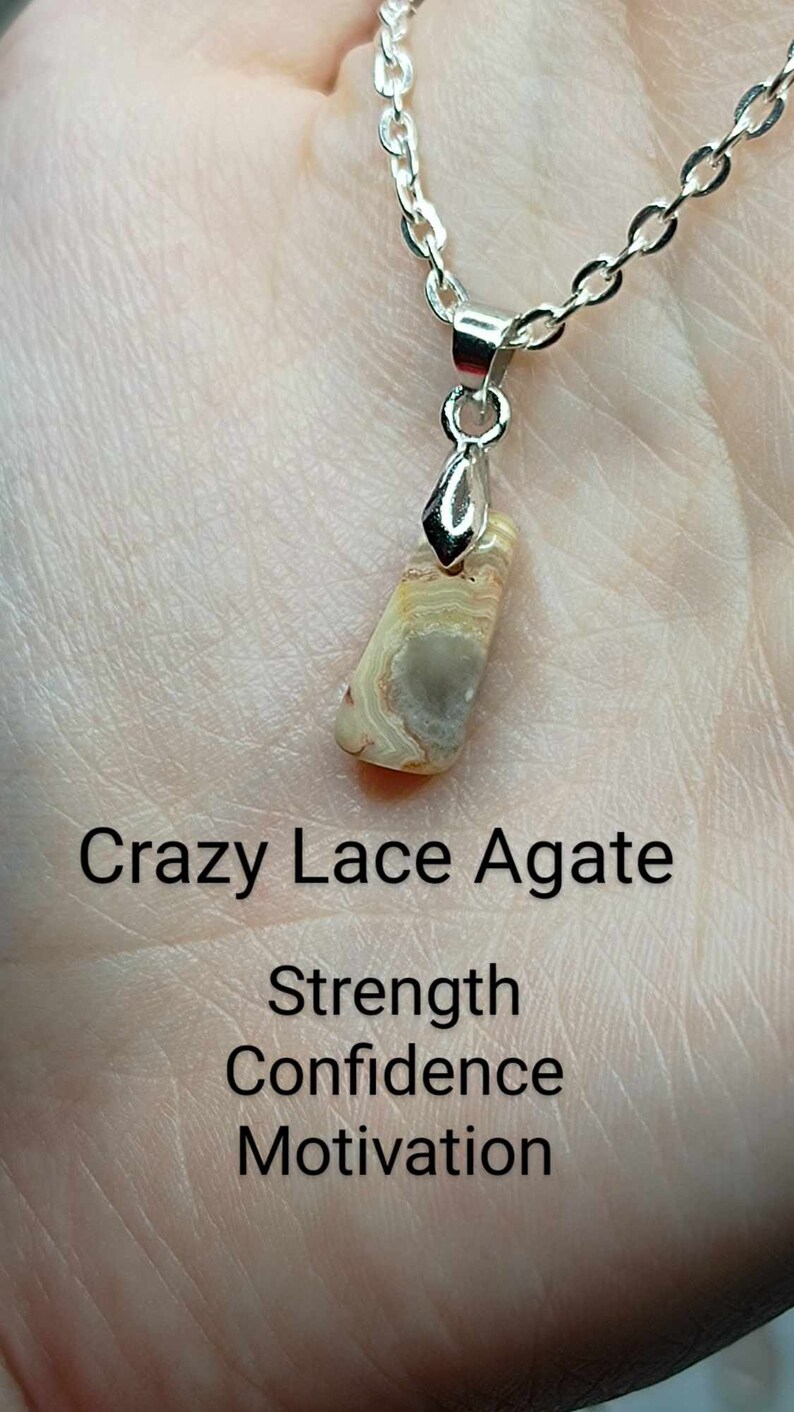 May include: A silver chain necklace with a pendant made of a light brown and white Crazy Lace Agate stone. The text "Strength Confidence Motivation" is written below the stone.