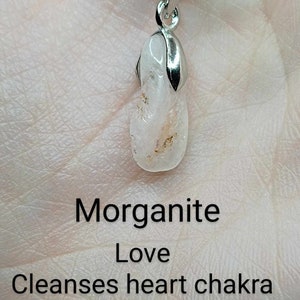 May include: A silver chain necklace with a pendant made of a clear, pink morganite crystal. The pendant is shaped like a teardrop and has a silver bail. The text "Morganite Love Cleanses heart chakra Peace" is written below the pendant.