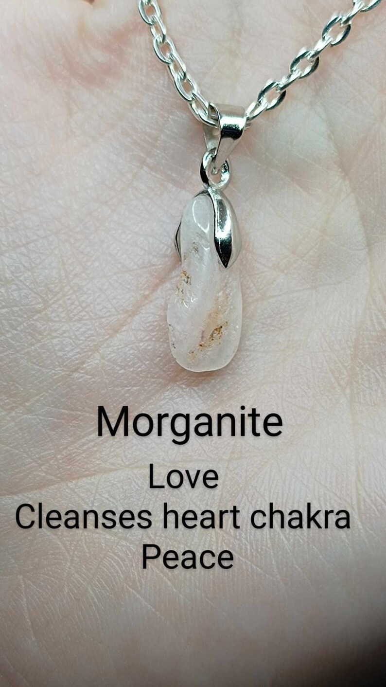 May include: A silver chain necklace with a pendant made of a clear, pink morganite crystal. The pendant is shaped like a teardrop and has a silver bail. The text "Morganite Love Cleanses heart chakra Peace" is written below the pendant.