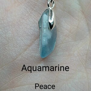May include: A silver chain necklace with a pendant featuring a light blue aquamarine stone. The text "Aquamarine" is below the stone, and the text "Peace Inspiration Growth" is below that.