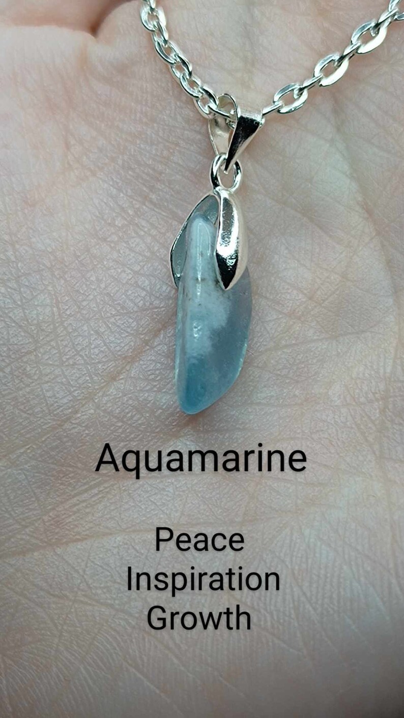 May include: A silver chain necklace with a pendant featuring a light blue aquamarine stone. The text "Aquamarine" is below the stone, and the text "Peace Inspiration Growth" is below that.